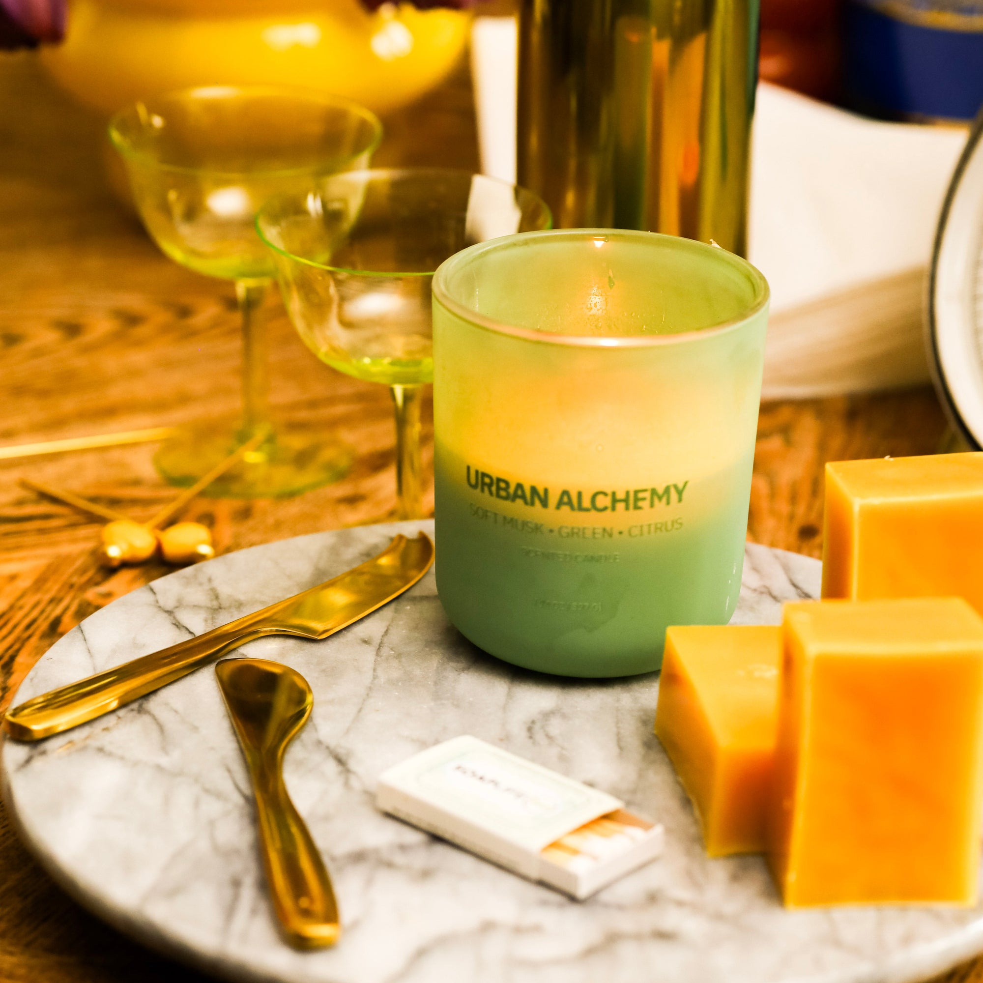 Candle labeled 'Urban Alchemy' on a marble surface with cheese and wine glasses in the background