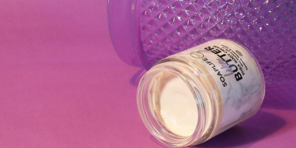 SoapLife360 Shea Body Butter in a clear glass jar lying on its side against a purple background with a textured purple bottle behind it