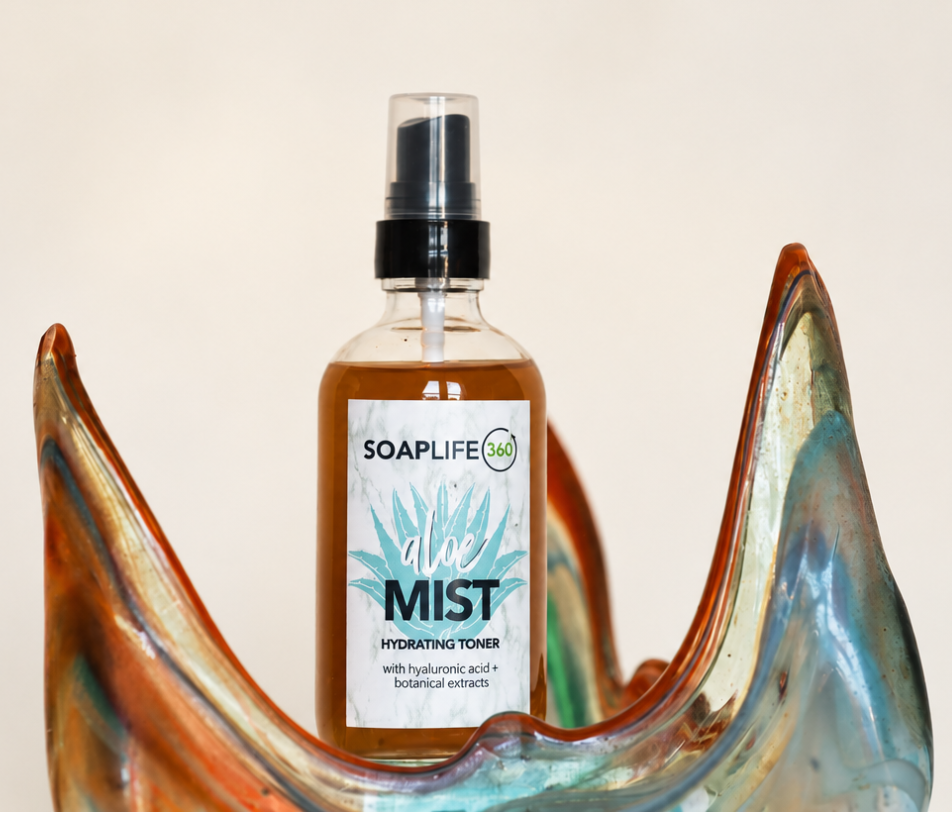 A bottle of Soaplife360 Aloe Mist hydrating toner with a clear spray top, placed in front of a decorative, curved glass dish with orange, blue, and green tones against a plain off-white background.