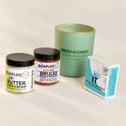 Four skincare and body care products arranged on a light beige studio background: a large mint green candle jar labeled Urban Alchemy, a Soaplife Shea Cream jar, a Soaplife Body Polish jar, and a blue-and-white face it  soap box.