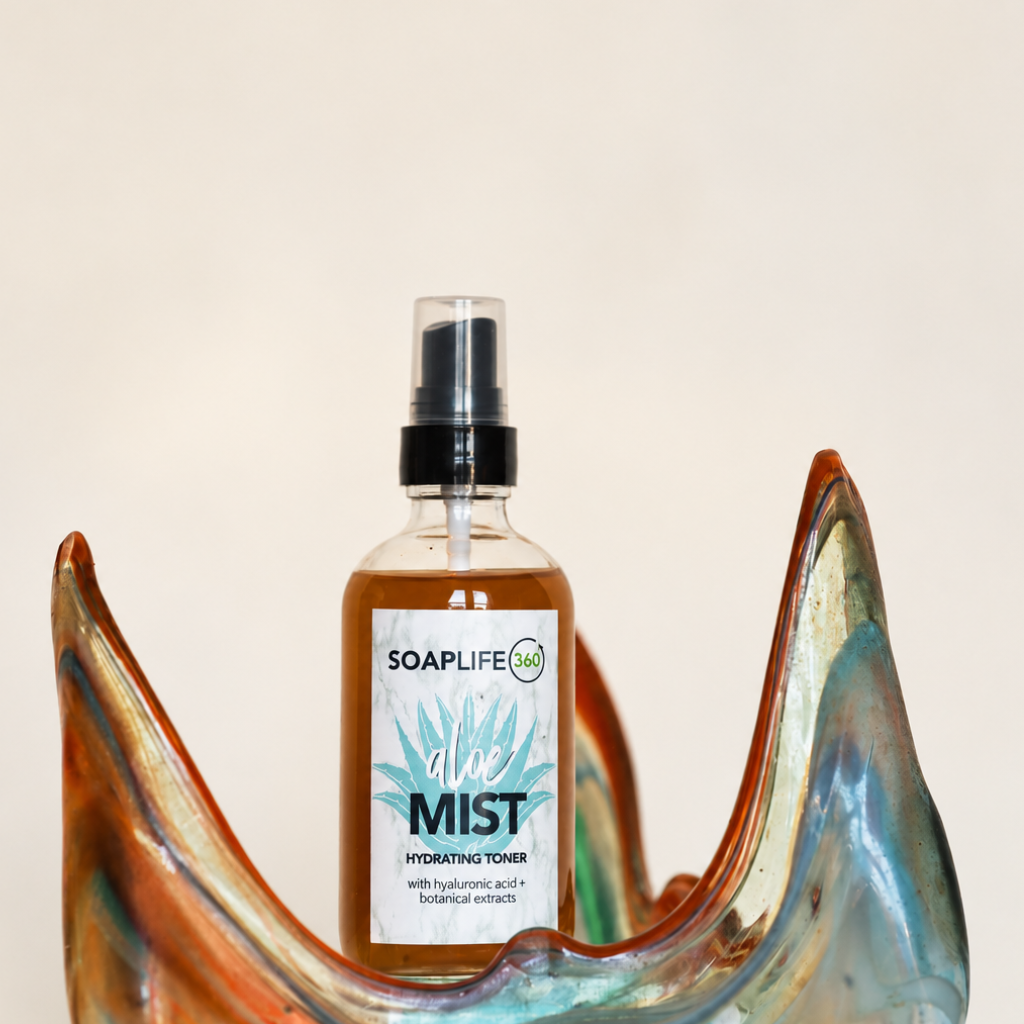 A bottle of Soaplife360 Mist hydrating toner with a clear spray top, placed in front of a decorative, curved glass dish with orange, blue, and green tones against a plain off-white background.