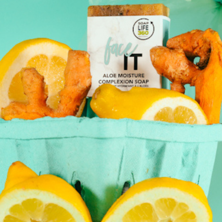 A gift set in a light blue paper basket featuring a box of face IT Aloe Moisture Complexion Soap by SoapLife360, surrounded by fresh lemon slices and orange ginger roots, set against a solid mint green background.