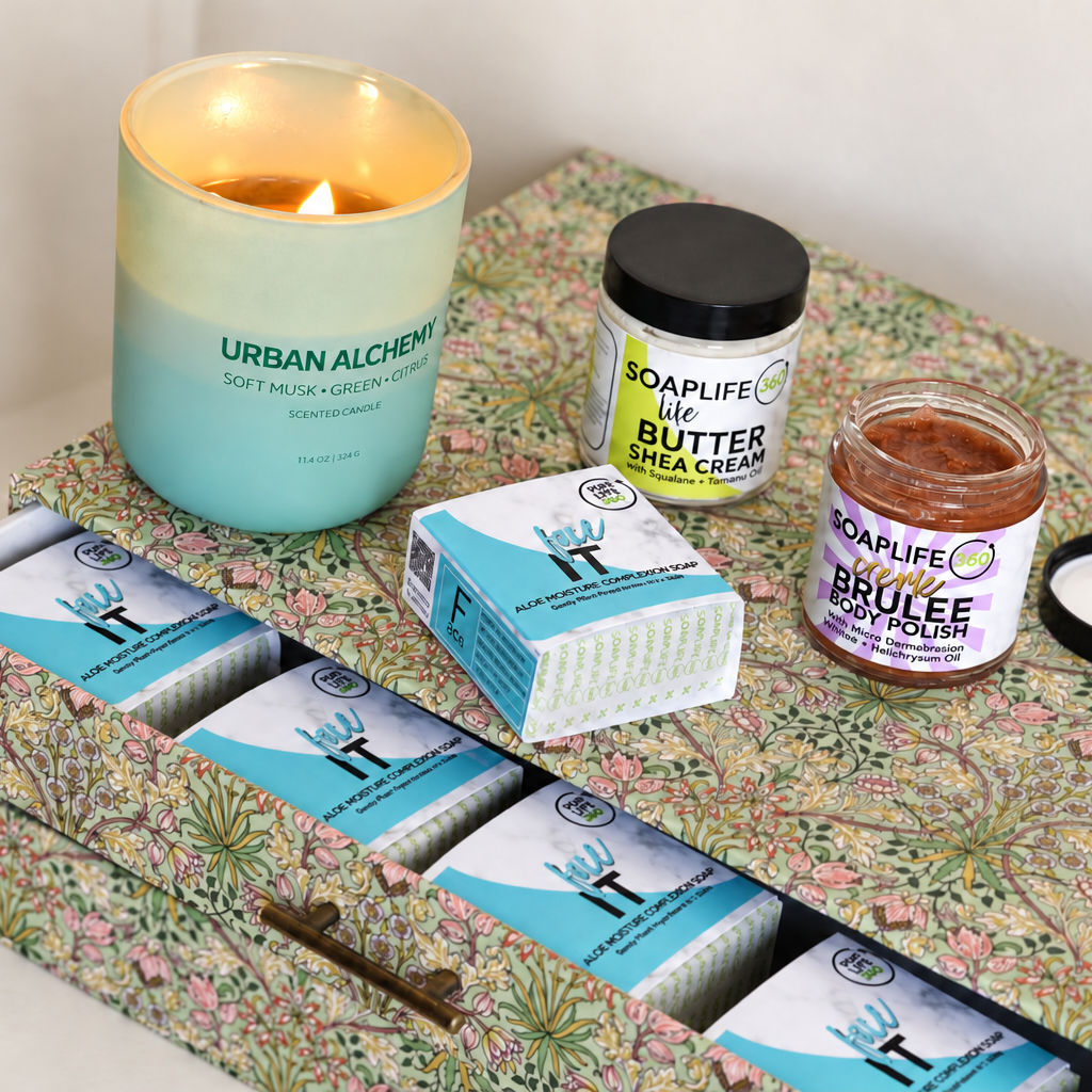 A curated self-care gift set arranged on a floral-patterned box. The set includes a lit Urban Alchemy beeswax soy candle, a jar of shea cream butter stick, a jar of creme brulee body polish